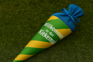 Fotografie einer bunten Zuckertüte mit der Aufschrift ‚Willkommen in der Kiekemal‘, liegend auf grünem Gras – symbolisch für den ersten Schultag.