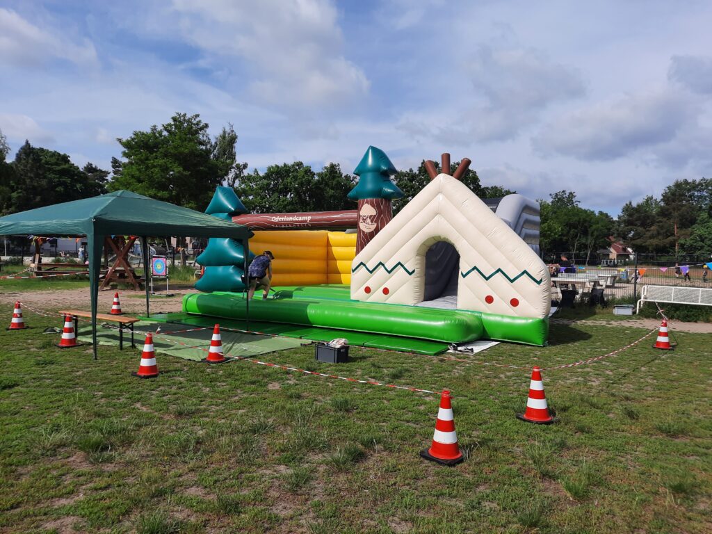 Aufblasbare Spielstation im Outdoor‑Bereich, gestaltet wie ein kleines Wald‑ und Camp‑Szenario, mit Zelt‑Elementen und Bäumen. Eine Person richtet die Station ein; im Hintergrund stehen ein Pavillon und Bäume.