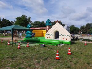 Aufblasbare Spielstation im Outdoor‑Bereich, gestaltet wie ein kleines Wald‑ und Camp‑Szenario, mit Zelt‑Elementen und Bäumen. Eine Person richtet die Station ein; im Hintergrund stehen ein Pavillon und Bäume.