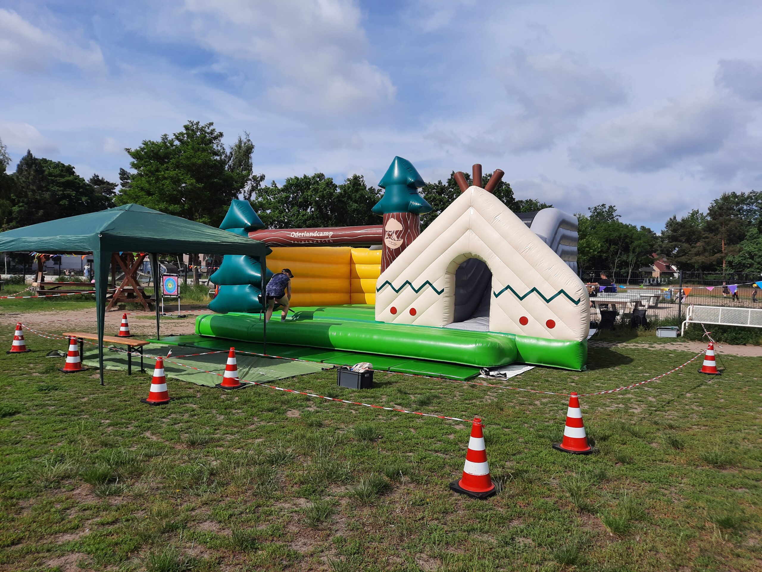 Aufblasbare Spielstation im Outdoor‑Bereich, gestaltet wie ein kleines Wald‑ und Camp‑Szenario, mit Zelt‑Elementen und Bäumen. Eine Person richtet die Station ein; im Hintergrund stehen ein Pavillon und Bäume.