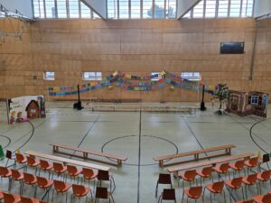 Dekorierte Turnhalle mit Bühne für eine Aufführung; bunte Namensgirlanden, Kulisse mit einem überdimensionalen Buch und einem Haus vor dem ein Zuckertütenbaum steht und Stuhlreihen für das Publikum.