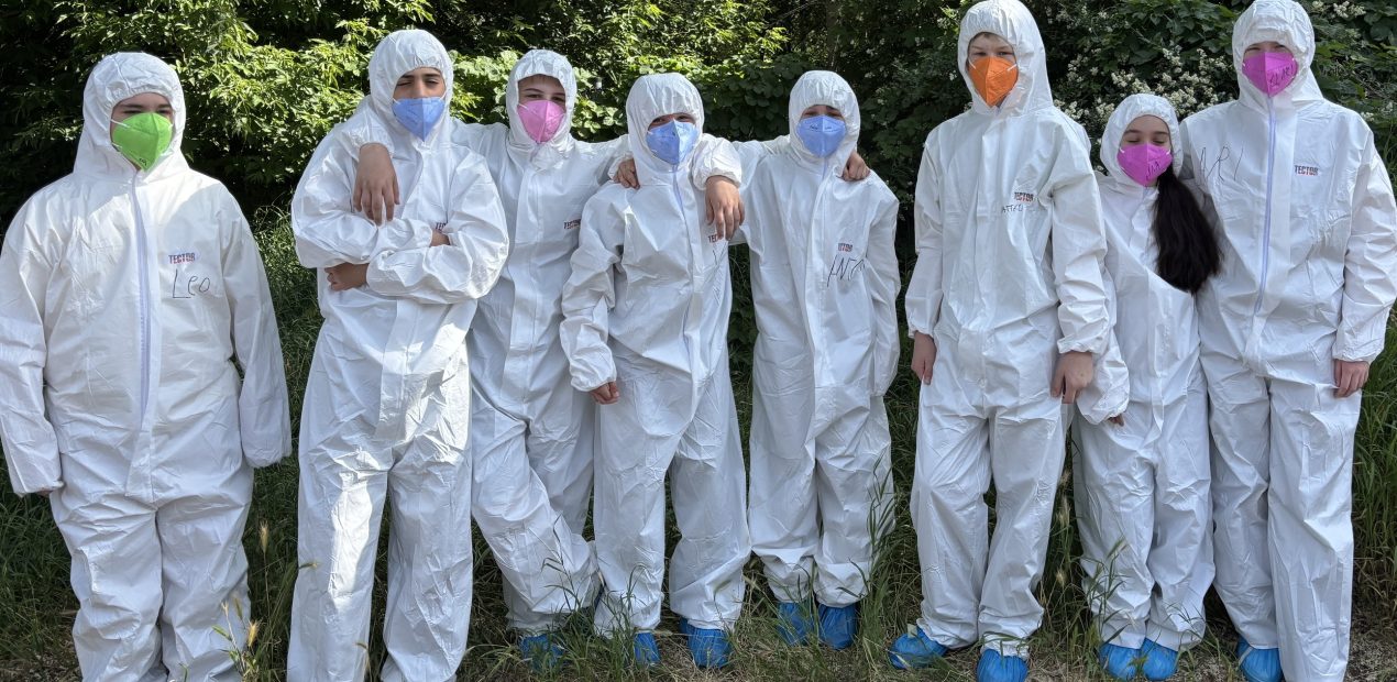 Gruppe von Kindern steht in weißen Schutzanzügen und farbigen Masken vor dichtem Grün.